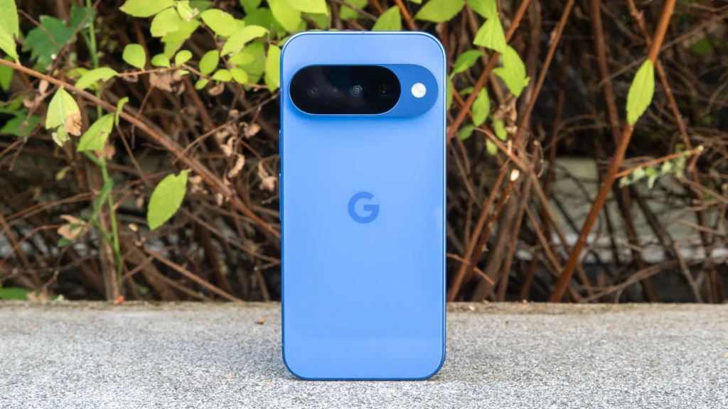 Google Pixel 10 smartphone in blue, showcasing the Google Pixel 10 specifications, standing on a concrete surface against a green leafy background.